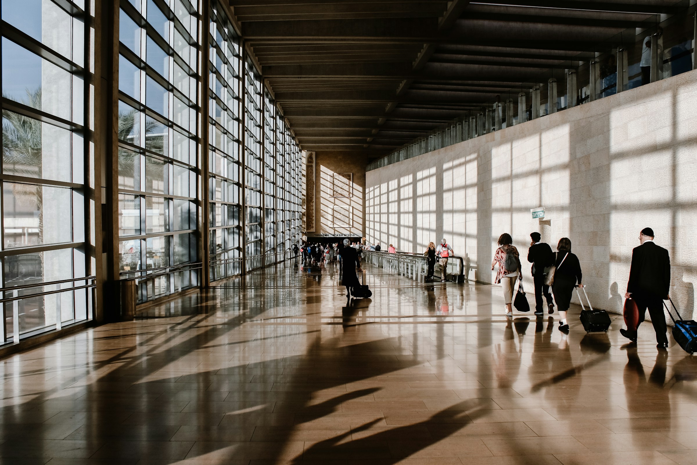 A spacious, modern airport terminal with large windows allowing natural light to flood the area. Several people are walking or standing, some with luggage. The floor is shiny and reflects the light and shadows from the windows. The ceiling has a series of beams running parallel to each other.
