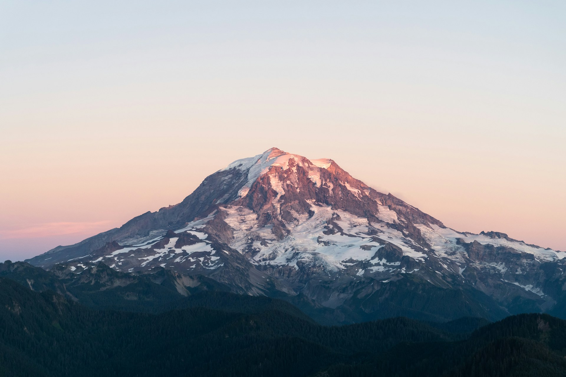 A majestic mountain peak covered in snow, with the top portion illuminated by the soft pink and orange hues of a setting or rising sun. The sky is clear with a gradient from light pink near the horizon to pale blue higher up. The lower slopes of the mountain are dark and rugged, contrasting with the bright, snow-covered summit.