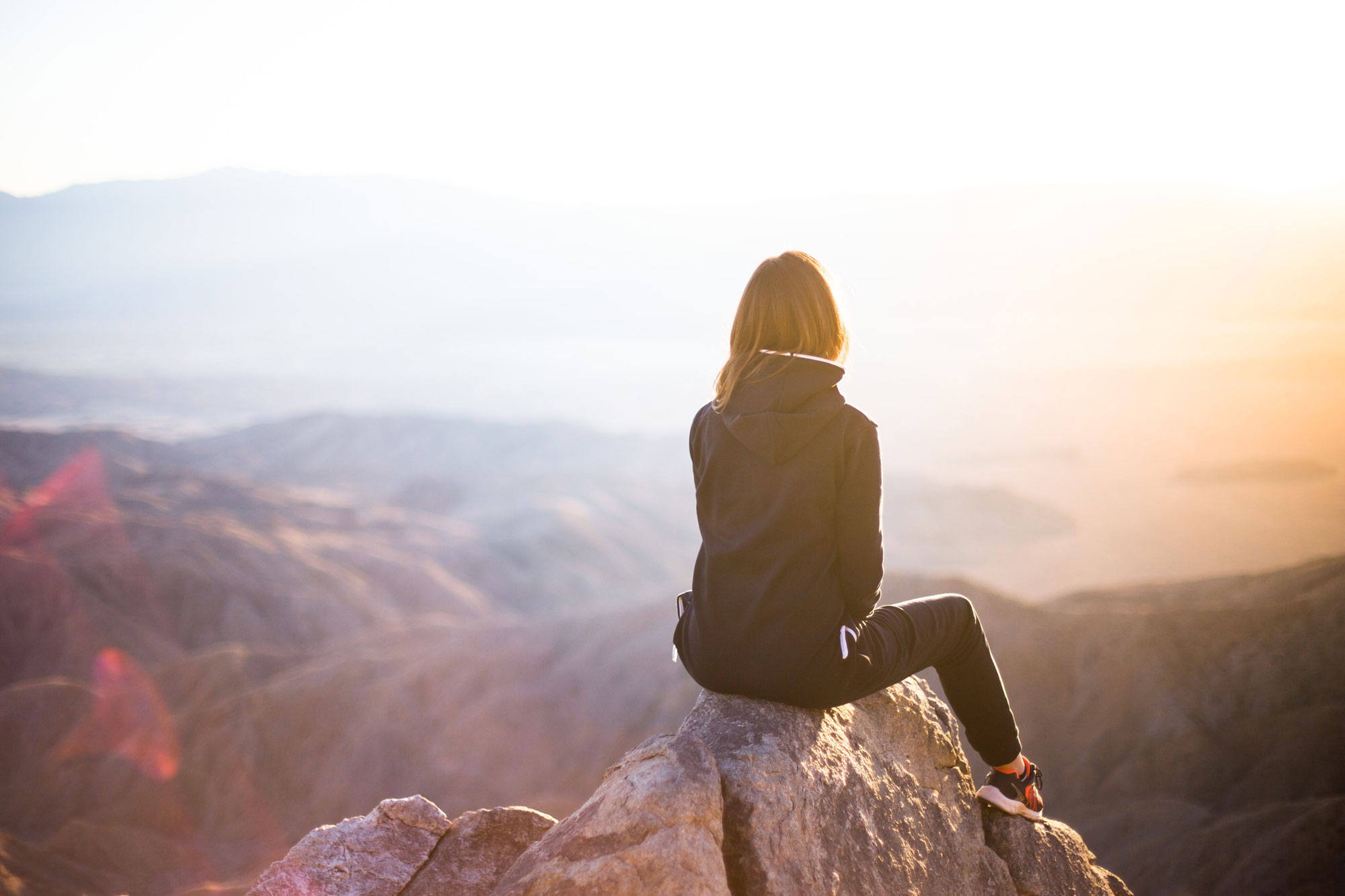 A person sitting on a rocky outcrop, overlooking a vast mountainous landscape during what appears to be sunrise or sunset. The person is wearing a dark jacket and pants, and the scene is bathed in warm, golden light.